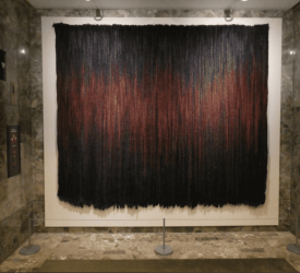 Large wall-mounted textile artwork featuring vertical dark fibers blended with deep red and orange tones, displayed in a museum setting.